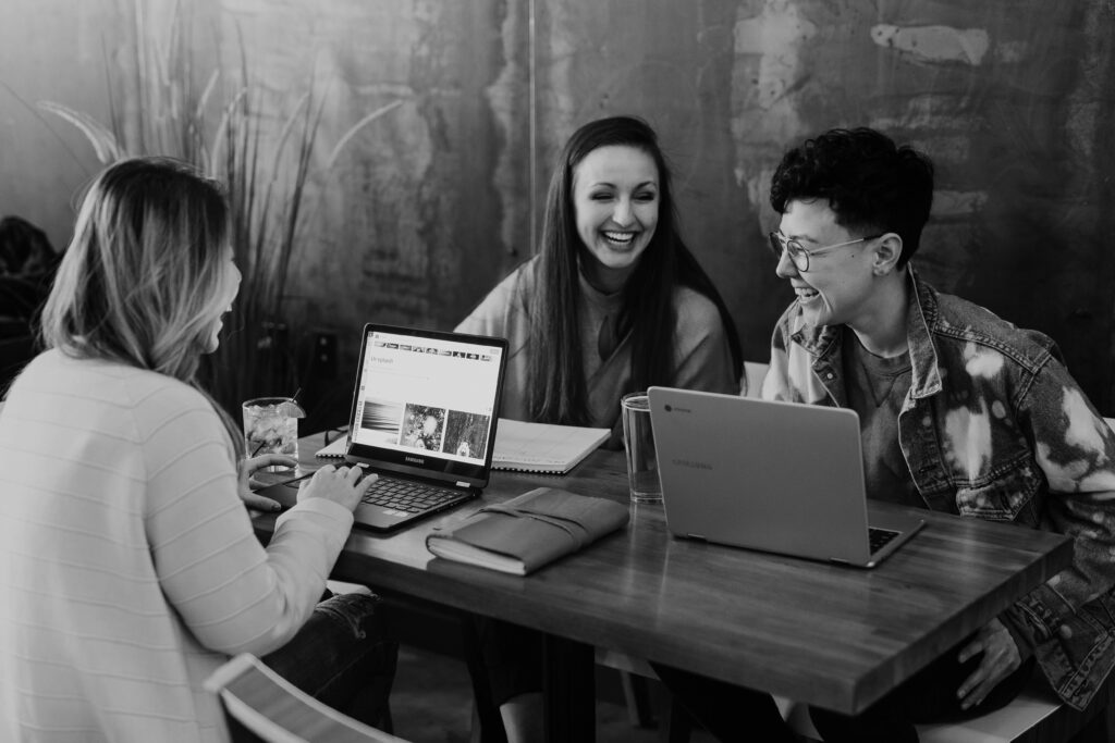 Training für gewaltfreie Kommunikation. schwarz-weiß Foto von drei Frauen, die mit Laptops an einem Tisch sitzen und lachen